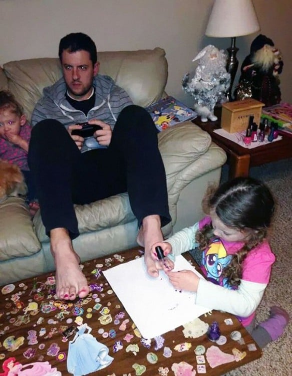 hombre sentado en un sillón jugando videojuegos mientras su hija le pinta las uñas 