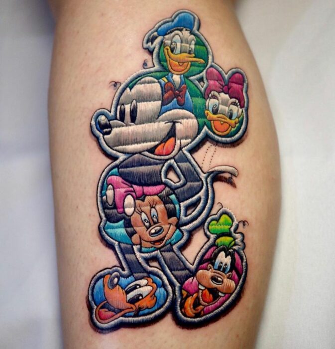 Tatuaje que imita a un parche cocido con mickey mouse compuesto por las imágenes de dondald duck daisy duck minnie mouse pluto el perro y goofy pato donald la pata daisy el ratón miguelito la ratona minnie pluto el perro y tribilín