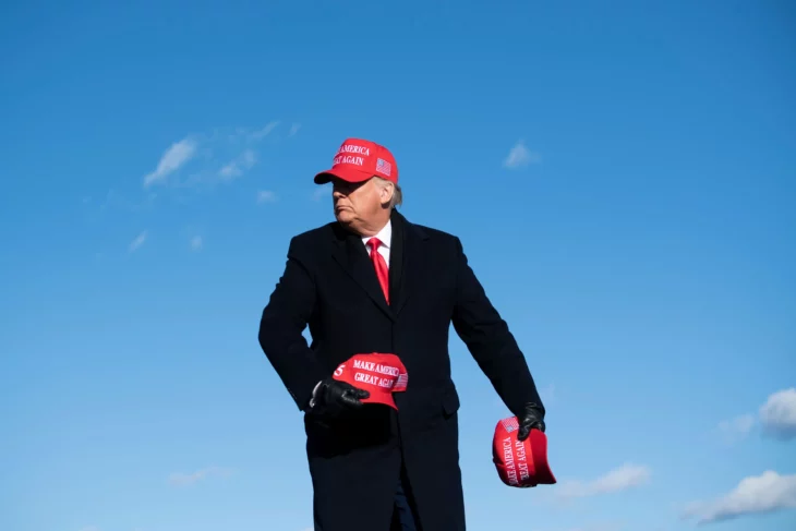 Donald trump con una gorra roja de haz grande nuevamente a Estados Unidos Make Aerica Great again con traje negro guantes negros y corbata roja sostiene dos gorras rojas en sus manos