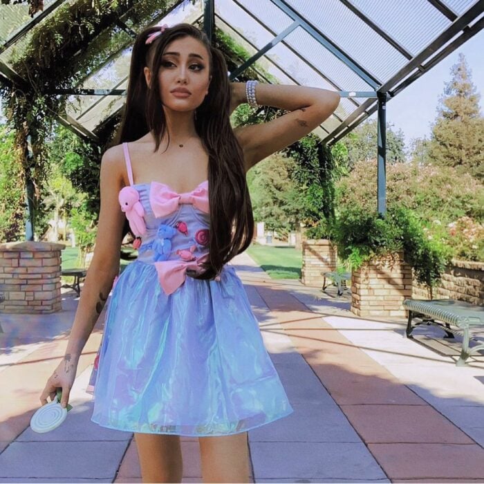 Paige Nieman con vestido azul que tiene peluches y moños rosas en un patio al aire libre con el pelo en coletas y con una mano en su nuca