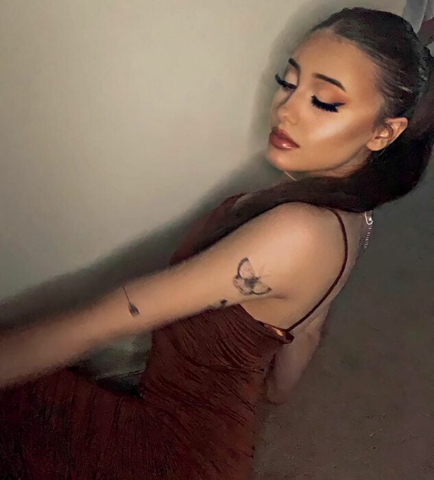 Paige Niemann con vestido de tirantes café mirando hacia abajo con los ojos casi cerrados con coleta y luciendo el tatuaje de mariposa en su brazo izquierdo