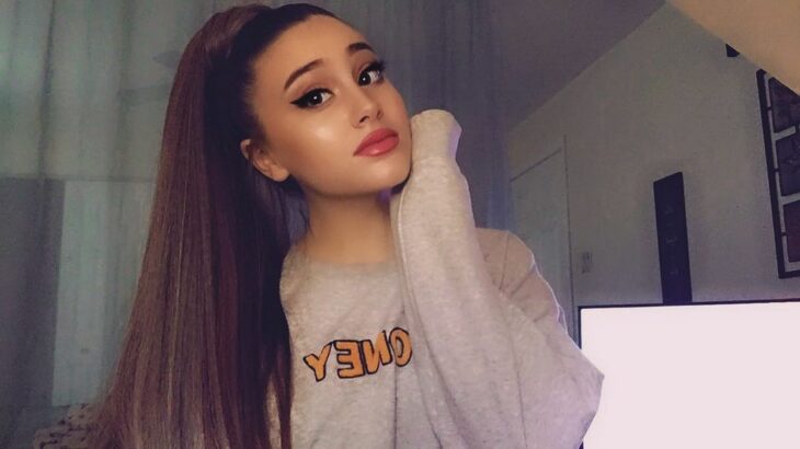 Paige Niemann en una sudadera gris tocándose la mejilla con la manga y usando una coleta como la de Ariana Grande