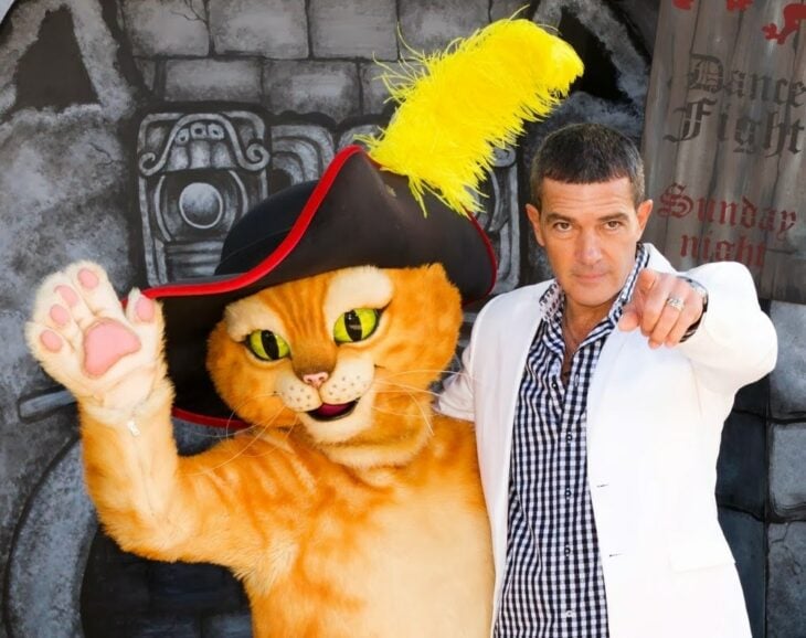 Antonio banderas vestido de blanco con camiza a cuadros en la premier del gato con botas y el último deseo junto a una botarga del titular gato con botas