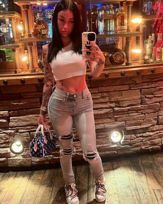 Bhad Bhabie tomando selfie top de tanque blanco pantalones de mezclilla rotos y bolso de mano en un club antro con piso de madera luces de noche bebidas alcohólicas