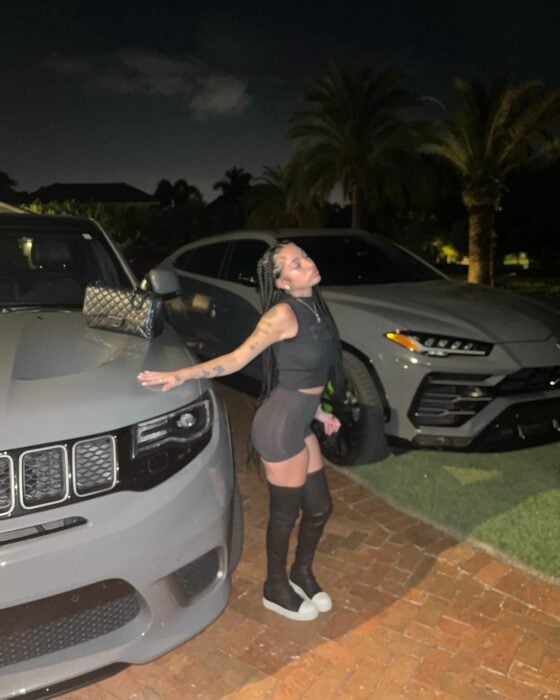 Bhad Bhabie Danielle en el estacionamiento con pantalones cortos y medias a medio muslo negras zapatos blancos con mano sobre camioneta