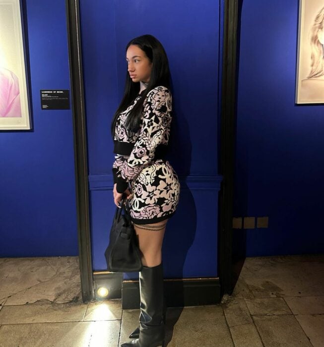 Bhad Bhabie de Perfil con bolso vestido negro blanco botas altas negras en cuarto azul exposición de arte