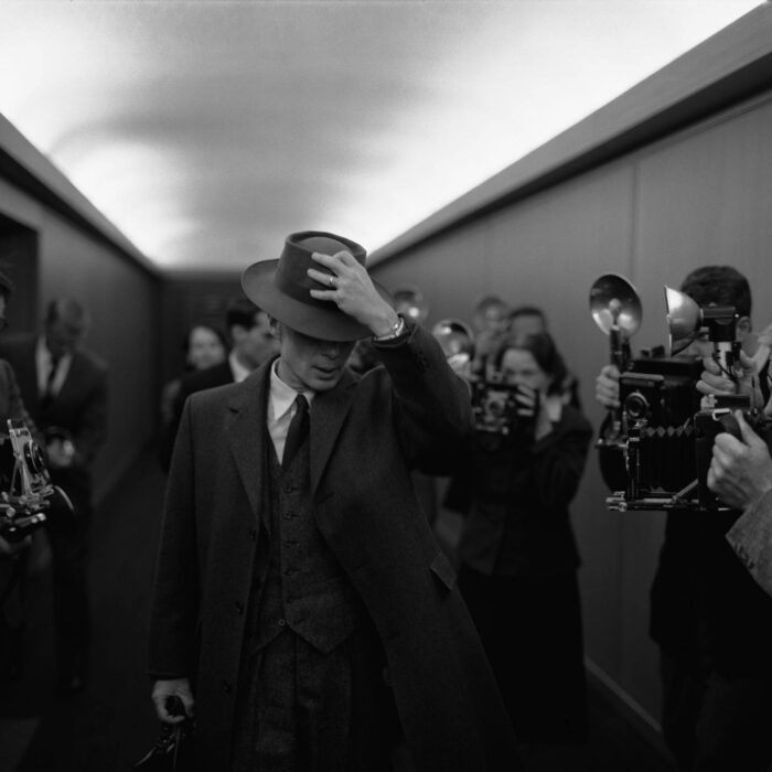 Imagen de Cillian Murphy interpretando a Robert Oppenheimer siendo seguido por un pasillo por periodistas con cámaras fotográficas