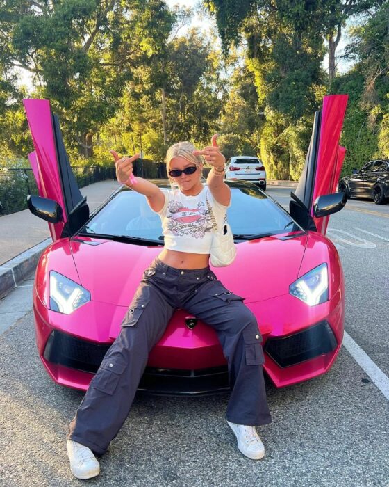 KAtie Sigmond sobre el cofre de un automóvil rosa con pantalones negros tenis y top blanco