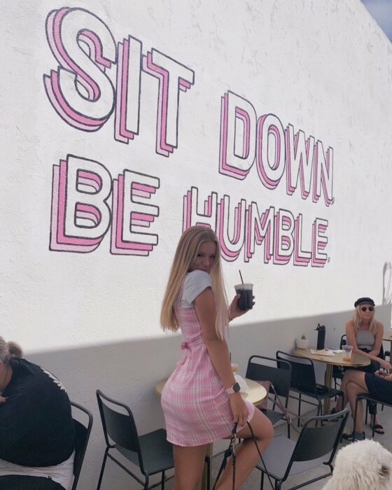 tiktoker tiktok influencer instagram Katie Sigmond en vestido verde apoyándose en un palo de golf tenis blancos en una cafetería vestida de rosa y blanco de espaldas con la leyenda sit down be humble siéntate sé humilde al fondo