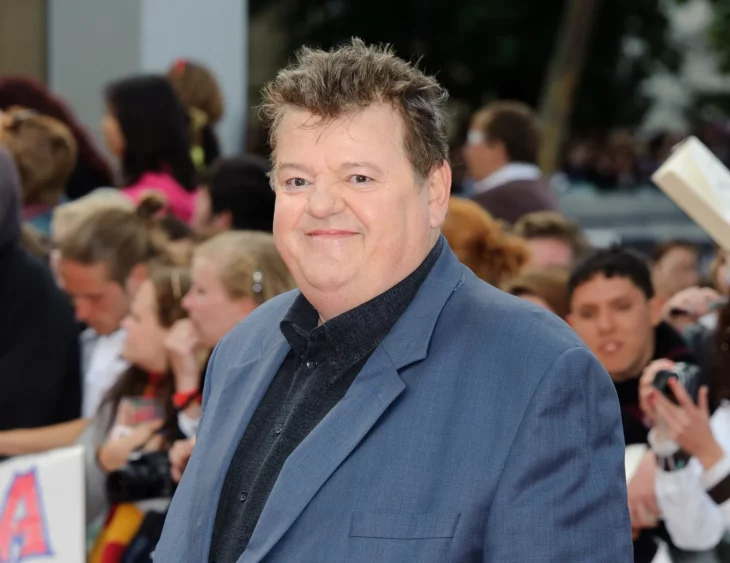 Robbie Coltrane en una presentación de traje azul con camisa negra sonriendo