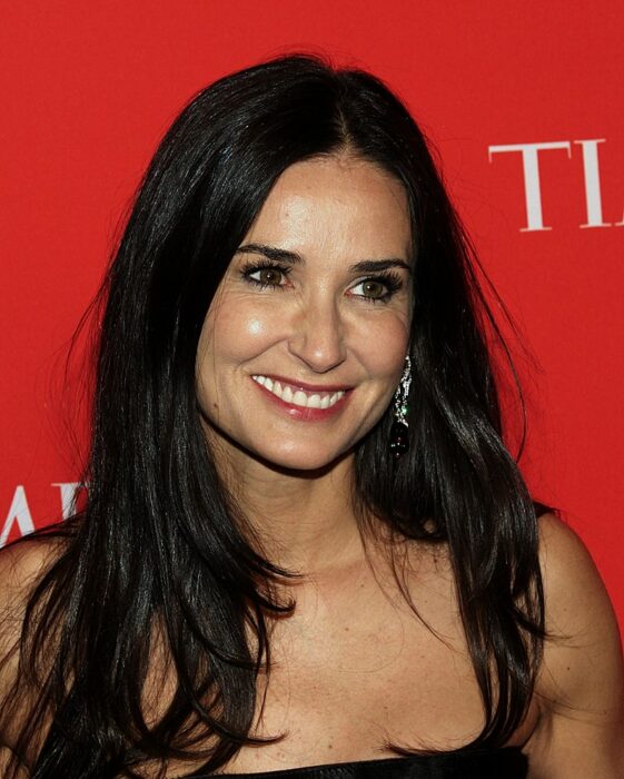 Demi Moore sonriendo cabello largo vestido negro frente a fondo rojo