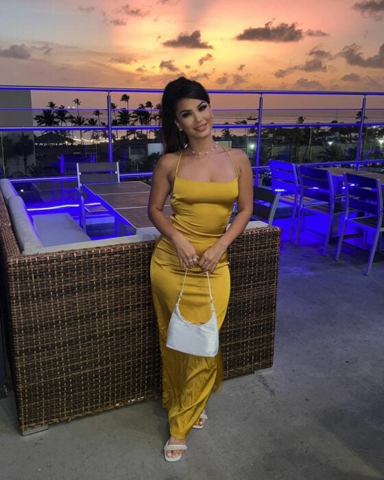 Shye Lee Vestido amarillo bolso blanco frente al atardecer en terraza de restaurante